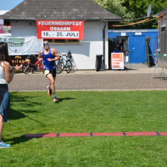 Ein Mann läuft auf einem Rasenfeld und trägt eine Sonnenbrille und ein blaues Tanktop. Eine Frau steht in der Nähe und beobachtet ihn. Dahinter befindet sich ein Gebäude mit einem Schild, auf dem 'Feuerwehrfest Ossarn 18. - 20. Juli' steht.
