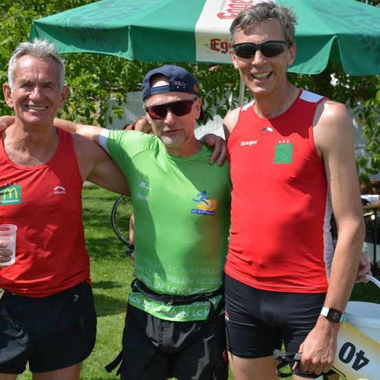 Drei Männer in Sportkleidung lächeln und posieren für ein Foto unter einem grünen Regenschirm. Einer trägt ein McDonald's-Shirt, ein anderer ein grünes Shirt mit weißem Text und der dritte ein rotes Shirt mit einem grünen Aufkleber.