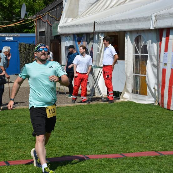 Ein Mann in einem hellblauen T-Shirt und schwarzen Shorts läuft auf einem Rasenfeld mit einem Nummer-113-Tag. Hinter ihm stehen zwei Männer in weißen Hemden und roten Hosen in der Nähe eines Zeltes.