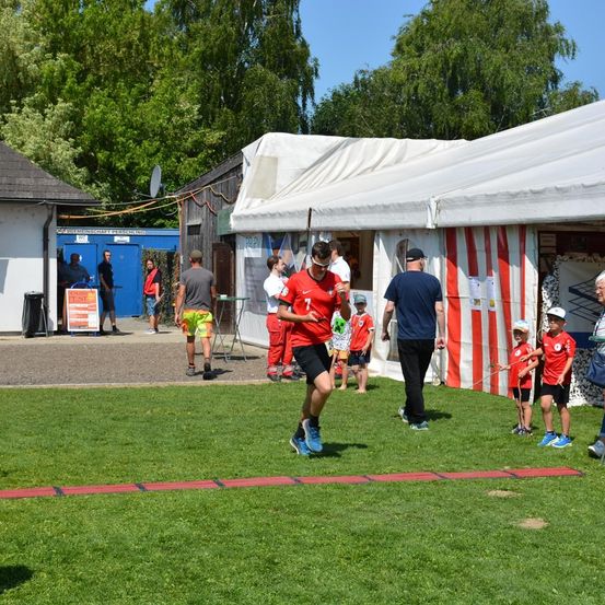 Mehrere Personen befinden sich auf einem Rasenfeld, wobei ein Mann auf einer roten Linie läuft. Mehrere Zelte und Gebäude sind im Hintergrund. Einige Menschen stehen in der Nähe der Zelte und Gebäude.