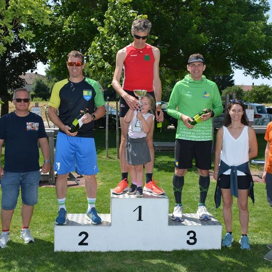 Sechs Personen stehen auf einem Podium, jede hält eine Flasche. Sie feiern wahrscheinlich einen Sieg. Das Podium hat die Zahlen 1, 2 und 3 darauf. Der Hintergrund besteht aus Grün und geparkten Autos.