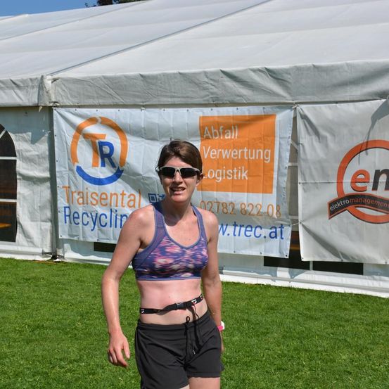 Eine Frau in einem Sport-BH und Shorts steht vor einem weißen Zelt, lächelt und posiert für ein Foto. Hinter ihr befinden sich zwei Banner, eines mit einem orangefarbenen Logo und dem Text 'Abfall Verwertung Logistik' und ein anderes mit einem Logo und dem Text 'Elektromanagement'.