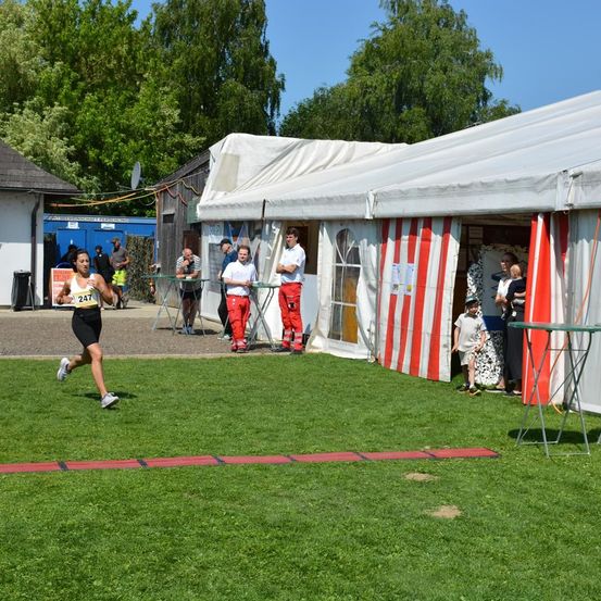 Ein Läufer mit der Nummer 247 sprintet auf einem Rasenfeld, mit Zelten und Leuten in der Nähe. Im Hintergrund stehen Bäume und ein Gebäude.