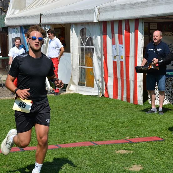 Ein Mann läuft auf einem Rasenfeld mit der Nummer 46 auf seinem Shirt. Hinter ihm hält ein Mann mit Hut eine Trompete. Es gibt Zelte mit Menschen in der Umgebung.