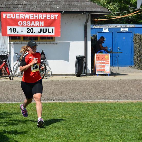 Eine Frau läuft einen Marathon in einem roten Trikot. Die Veranstaltung wird von der örtlichen Feuerwehr gesponsert. Ein Schild zeigt die Veranstaltungstermine an.