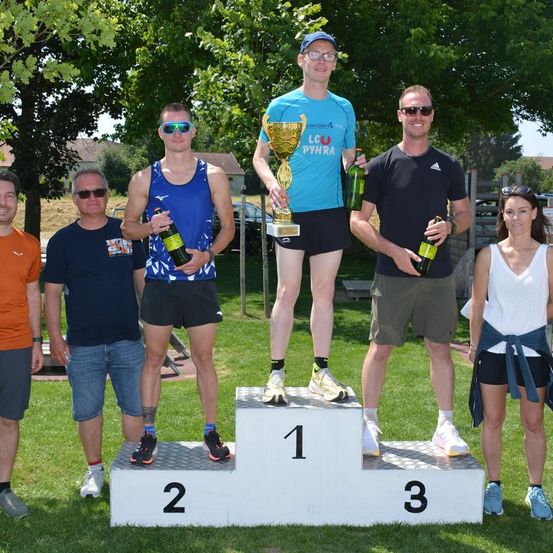 Eine Gruppe von Menschen steht auf einem Podest, einer hält eine Trophäe. Der Gewinner des ersten Platzes hält eine Trophäe und eine Flasche, der Gewinner des zweiten Platzes hält eine Flasche und der Gewinner des dritten Platzes hält eine Flasche. Bäume und ein Haus sind im Hintergrund.