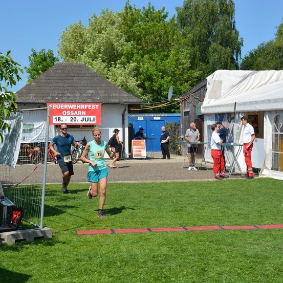 Ein Außenbereich mit einem Grasfeld, einem roten Banner und mehreren Personen, darunter eine Frau, die mit der Nummer 19 läuft, während andere stehen, gehen oder sich in der Nähe eines Zeltes befinden.