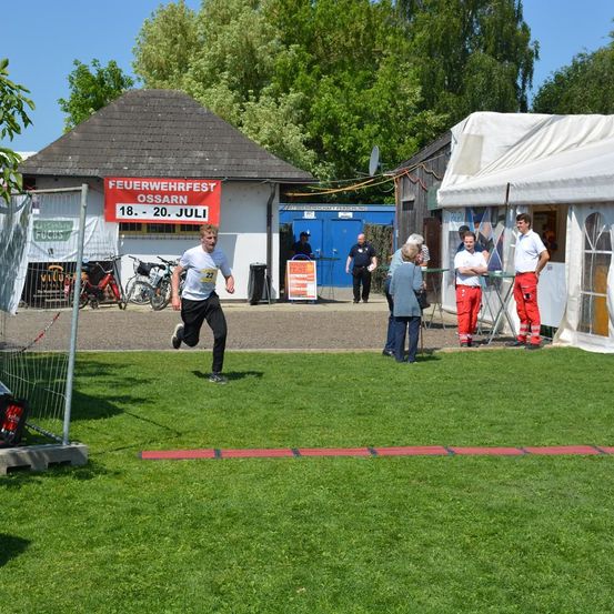 Ein Läufer sprintet auf einem Rasenfeld und trägt ein weißes Shirt und schwarze Hose. Hinter ihm stehen mehrere Personen in der Nähe eines Gebäudes mit einem roten Schild, das Feuerwehrfest Ossarn besagt.