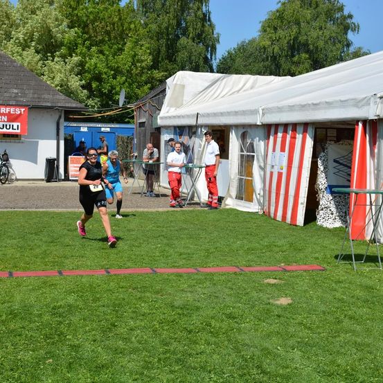 Eine Frau mit der Nummer 29 läuft auf einem Rasenfeld und überquert eine rote Linie. Hinter ihr stehen zwei Personen vor einem weißen Zelt. Einer von ihnen trägt einen roten Overall. Es gibt Leute, die in der Nähe des Zeltes stehen. Bäume und ein Haus sind im Hintergrund.