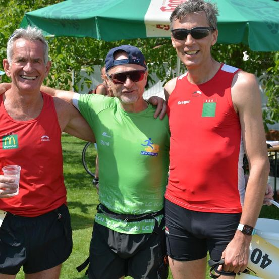 Drei Männer in Sportbekleidung posieren für ein Foto. Der linke Mann hält ein Getränk, der mittlere trägt einen blauen Hut und der rechte Mann hat ein rotes Shirt an. Sie stehen unter einem grünen Regenschirm.