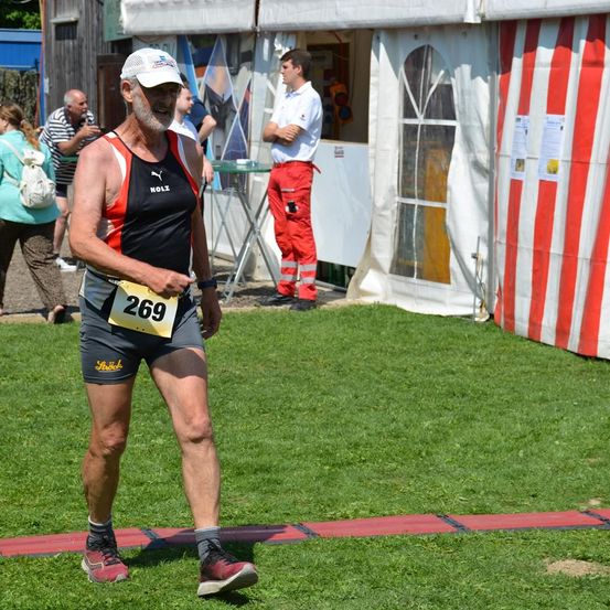 Ein älterer Mann mit einer weißen Kappe läuft auf einem Rasenfeld, trägt ein schwarz-rotes Shirt mit der Nummer 269. Dahinter befinden sich Zelte und ein Mann in reflektierender Kleidung.