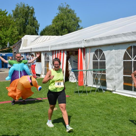 Eine Frau läuft auf einem Rasenfeld und trägt ein grünes Shirt mit der Nummer 41. Hinter ihr winkt ein Mann in einem bunten Kostüm. In der Nähe steht ein Zelt mit Tischen und Stühlen.
