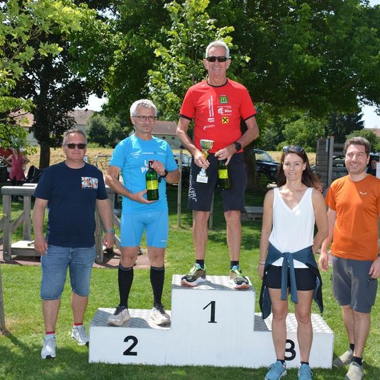 Eine Gruppe von Menschen steht auf einem Podest mit der Nummer 1. Ein Mann hält einen Pokal, und ein anderer eine Flasche. Eine Frau steht neben dem Mann mit dem Pokal. Dahinter sind Bäume und geparkte Autos.