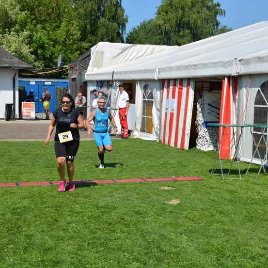 Zwei Läufer joggen auf einem Rasenfeld mit einem roten Hindernisparcours. Zelte und ein Gebäude sind im Hintergrund.