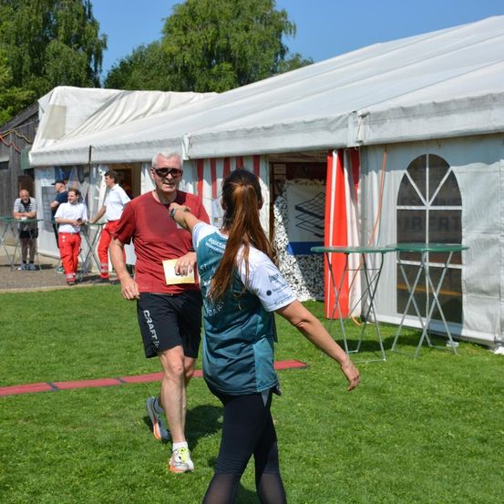 Ein Mann in Rot joggt an einer Frau mit einer Karte vorbei auf einem Rasenfeld mit Zelten und Tischen im Hintergrund.