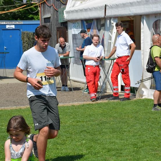 Ein Marathonläufer mit der Nummer 114 auf seinem Shirt joggt auf dem Rasen. In der Nähe stehen mehrere Personen um einen Tisch und führen möglicherweise ein Gespräch. Im Hintergrund befinden sich einige Zelte.