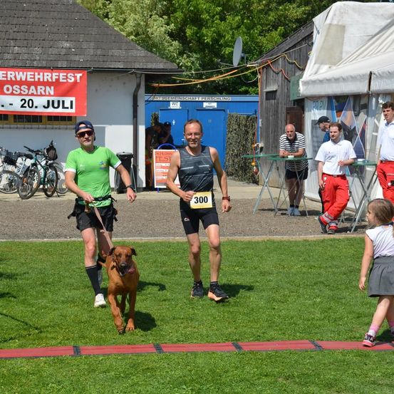 Zwei Männer joggen mit einem Hund an der Leine im Park. Ein Mann trägt die Nummer 300 auf seinen Shorts. Ein Banner steht 'Ossarnerwehrfest'. Dahinter stehen Menschen in der Nähe eines weißen Zeltes.