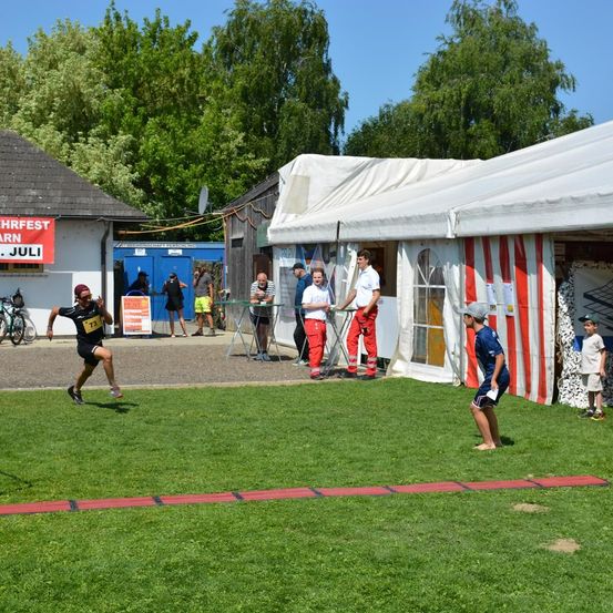 Eine Gruppe von Menschen ist im Freien in der Nähe eines Gebäudes. Eine Person läuft, während andere stehen oder gehen. Es gibt Zelte, Tische und Fahrräder.