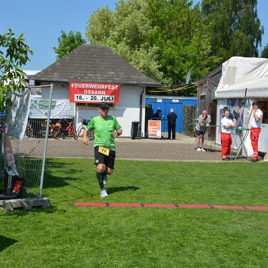 Ein Läufer joggt auf einem Rasenfeld. Ein rotes Banner mit den Worten 'Feuerwehrfest Ossarn' hängt an einem Gebäude. Andere Leute stehen in der Nähe eines Zeltes.