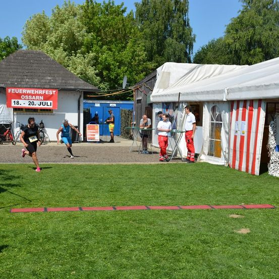 Läufer nehmen an einem Marathon teil. Zuschauer stehen in der Nähe eines Zeltes und beobachten. Gebäude und Bäume sind im Hintergrund.