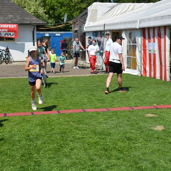 Ein junges Mädchen mit der Nummer 10 läuft auf einem Rasenfeld. Im Hintergrund steht ein Zelt. Leute stehen herum, einige tragen Helme, und ein Mann geht in der Nähe.