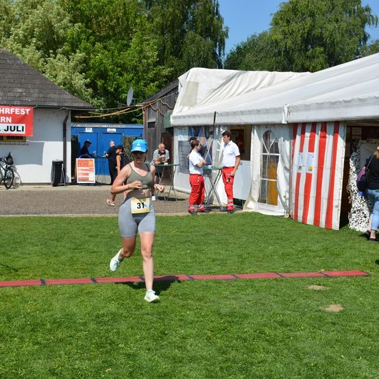 Eine Läuferin mit der Nummer 31 läuft auf einem Rasenfeld. Hinter ihr stehen mehrere Personen in der Nähe eines Zeltes und eines Gebäudes mit einem roten Schild.