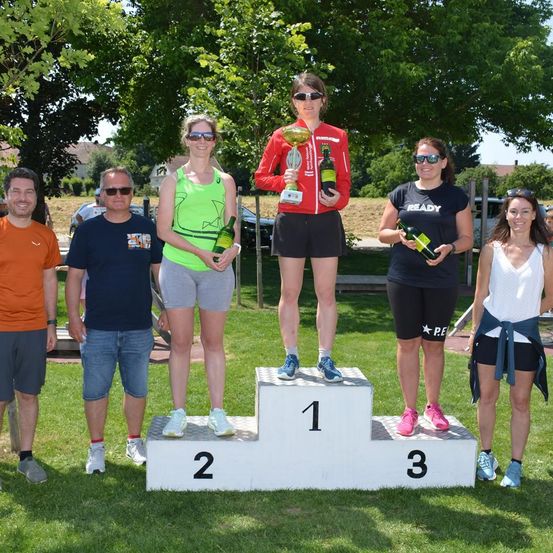 Eine Gruppe von Menschen steht auf einem Podest in einem Außenbereich, wobei eine Frau eine Trophäe und zwei Flaschen hält. Eine Frau in der Mitte hält eine Trophäe und eine weitere Frau hält eine Flasche. Ein paar Leute stehen neben ihnen, und ein Auto ist hinter ihnen geparkt. Bäume sind im Hintergrund.