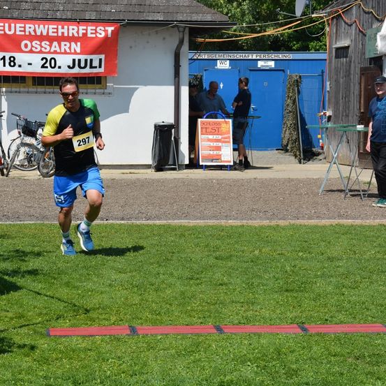 Ein Läufer mit der Startnummer 297 läuft auf einem Rasenfeld mit einer roten Leiter auf dem Boden. Hinter ihm befindet sich ein Gebäude mit einem Banner für das Feierwehrfest Ossarn.