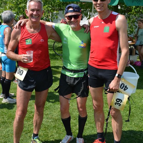 Drei Männer in Sportbekleidung stehen auf einem Rasenfeld und lächeln. Der Mann auf der linken Seite hält einen Becher, während der Mann in der Mitte eine Mütze trägt. Der Mann auf der rechten Seite trägt einen Eimer.