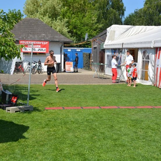 Ein Mann läuft auf einem Rasenfeld, hinter ihm steht ein Banner mit der Aufschrift Feuerwehrfest Osnarn 18. bis 20. Juli. Vor einem Zelt stehen Kinder und Erwachsene.