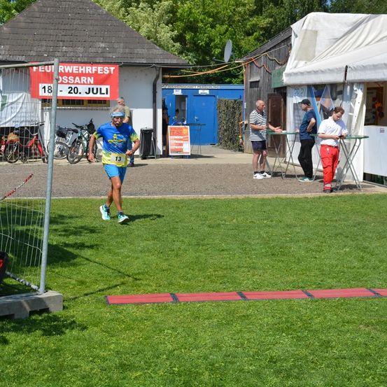 Ein Marathonläufer läuft an einem Banner vorbei, das für ein lokales Festival wirbt. Zwei Männer stehen in der Nähe eines Tisches, während ein anderer Mann in der Nähe geht. Fahrräder sind in der Nähe eines Gebäudes geparkt.