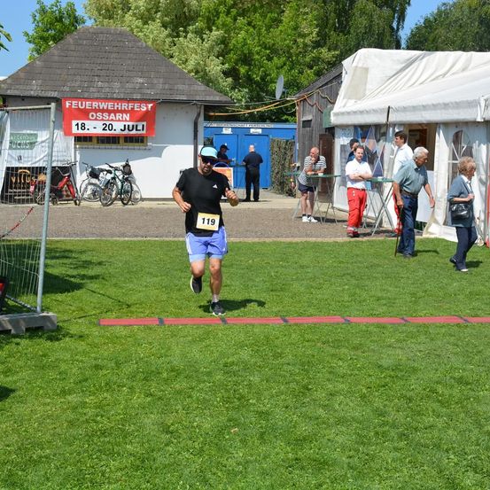 Ein Mann läuft auf einem Rasenfeld während des Feuerwehrfests in Ossarn, mit einem roten Schild darüber und Zuschauern und Gebäuden im Hintergrund.