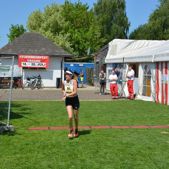 Eine Läuferin mit der Nummer 91 nimmt an einem Rennen auf einem Rasenfeld teil, mit Zelten und Gebäuden im Hintergrund.