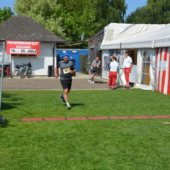 Ein Läufer mit blauer Mütze und Turnschuhen läuft auf einem Rasenfeld. Dahinter stehen mehrere Leute vor einem Zelt und einem Gebäude.