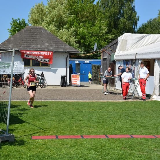 Eine Läuferin mit roter Kleidung und der Nummer 243 sprintet auf einem Rasenfeld. Hinter ihr befinden sich uniformierte Personen, ein Gebäude mit einem roten Banner und Fahrräder.