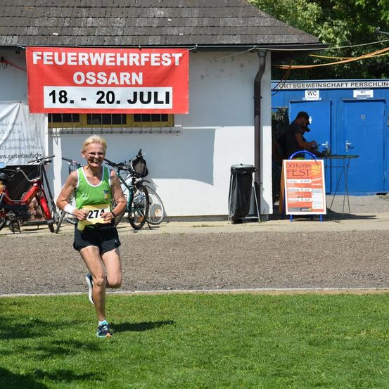 Eine Frau in einem grünen Trikot läuft einen Marathon in der Nähe eines Gebäudes mit einem Banner, das 'Feuerwehrfest Ossarn 18.-20. Juli' steht. Hinter ihr befinden sich Fahrräder und ein Mülleimer.