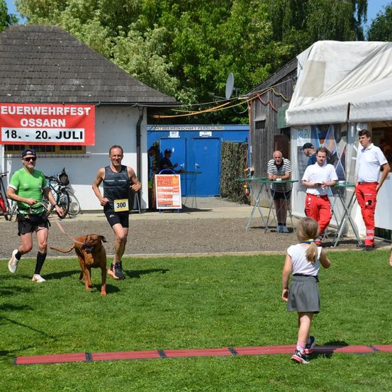 Zwei Männer rennen mit ihren Hunden auf einem Rasen. Dahinter befindet sich ein Gebäude mit einem roten Banner, auf dem Euerwehrfest Ossarn steht.