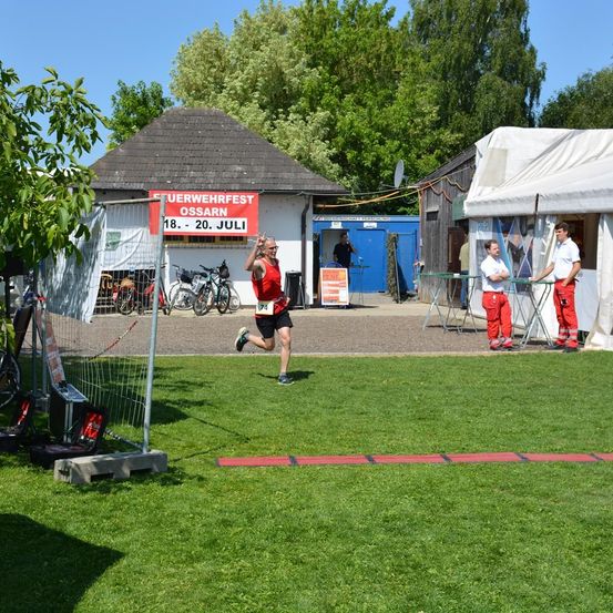 Ein Mann läuft in einem Rennen, in der Nähe eines Gebäudes mit einem Schild, das 'Uerwehrfest Ossarn 18. - 20. Juli' besagt. Zwei Männer in roten Overalls stehen vor einem Zelt.