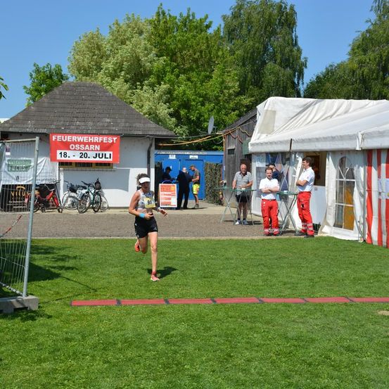 Eine Läuferin mit der Nummer 91 auf dem Trikot läuft auf einem Rasenfeld. Hinter ihr befinden sich Zelte und Fahrräder. Ein Banner mit der Aufschrift 'Feuerwehrfest Ossarn'. Mehrere Menschen stehen in der Nähe der Zelte.