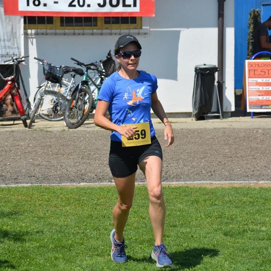Eine Läuferin in einem blauen Shirt und schwarzen Shorts mit der Nummer 59 auf ihrer Brust joggt vor einem weißen Gebäude. Hinter ihr stehen Fahrräder und ein Schild.