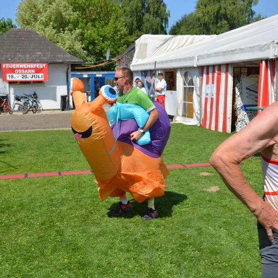 Ein Mann in einem riesigen Schneckenkostüm trägt ein Kind. Er befindet sich in einem Park. Hinter ihm steht ein Gebäude mit einem Schild, das Feuerwehrfest Ossarn anzeigt. Rechts ist ein weißes Zelt zu sehen.