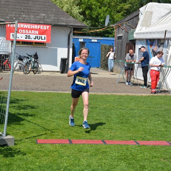 Eine Frau läuft bei einem Rennen mit der Nummer 262 auf ihrem Shirt. Ein Schild hinter ihr zeigt 'Erwerbsfest Ossarn 1-20. Juli'. Fahrräder sind in der Nähe des Gebäudes geparkt. Ein paar Leute stehen unter einem Zelt.