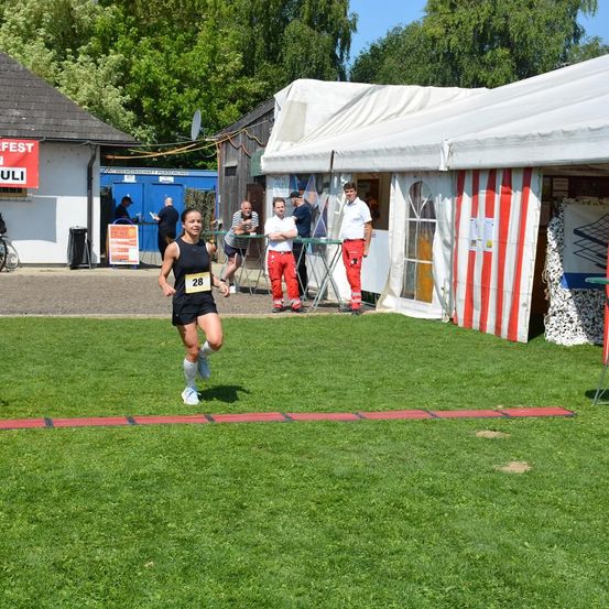 Eine Läuferin mit der Nummer 28 sprintet auf einem Rasenfeld. In der Nähe stehen zwei Männer in roten Anzügen bei einem Tisch. Dahinter ist ein Zelt mit einer rot-weiß gestreiften Wand aufgebaut.