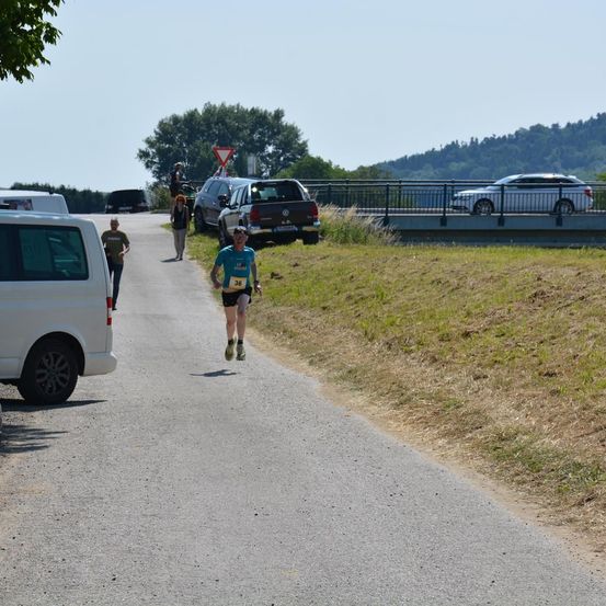 Ein Läufer mit blauem Shirt und der Nummer 36 joggt die Straße entlang, während zwei Personen in der Nähe gehen. Autos sind am Straßenrand geparkt, und eine Brücke ist im Hintergrund.