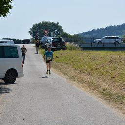 Ein Läufer mit blauem Shirt und der Nummer 36 joggt die Straße entlang, während zwei Personen in der Nähe gehen. Autos sind am Straßenrand geparkt, und eine Brücke ist im Hintergrund.