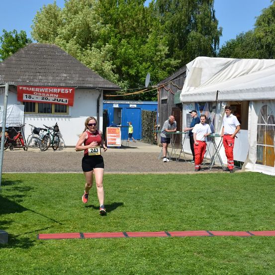 Eine Läuferin sprintet über eine Rasenfläche. Dahinter ist ein Gebäude mit einem Schild, auf dem 'Feuerwehrfest Ossarn' steht. Mehrere Menschen stehen herum, zwei in roten Overalls, einer in einem gestreiften Hemd.