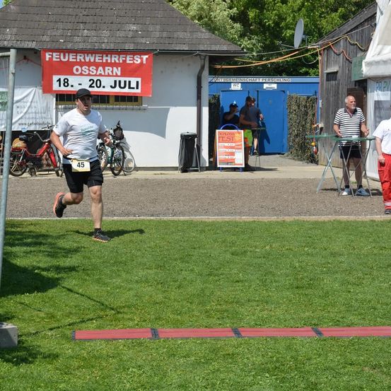 Ein Mann rennt auf einem Rasenfeld, mit einem roten Banner, das 'Feuerwehrfest Ossarn' steht, und einem Gebäude im Hintergrund. Mehrere Fahrräder sind in der Nähe des Gebäudes geparkt.