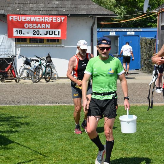 Zwei Männer gehen auf einem Rasenfeld, einer hält einen Eimer. Dahinter steht ein Gebäude mit einem roten Banner. Fahrräder sind davor geparkt. Ein Mann in einem weißen Shirt geht auf dem Bürgersteig.