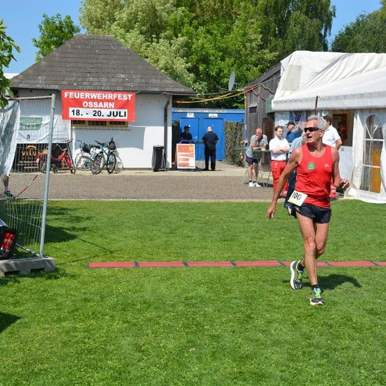 Ein Läufer in einem roten Shirt mit der Nummer 100 darauf läuft in einem Rennen. Hinter ihm stehen Menschen und schauen zu, und es gibt ein Zelt mit einem roten Banner.