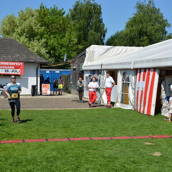 Ein Läufer bei einem Triathlon trägt ein nummeriertes Startnummerband und eine Sonnenbrille. Mehrere Personen stehen in der Nähe eines Zelts. Ein Gebäude mit einem Schild, das 'ERWERHFERST - 20. JULI' steht, ist im Hintergrund.